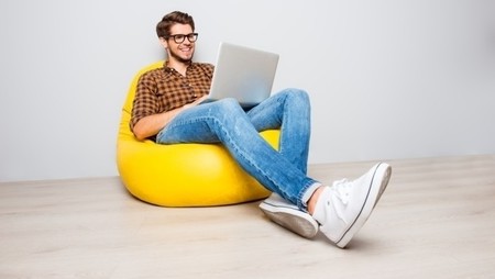 Happy young man sitting in yellow pouf and using laptop