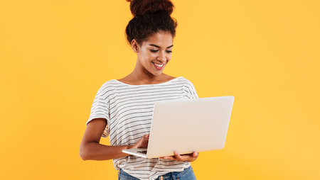 A smiling young woman holding a laptop and standing in front of a yellow background
